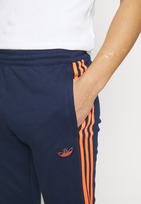 Tmavě modré teplákové kalhoty z bavlněné směsi s oranžovými bočními pruhy, elastickým pasem a předními kapsami; na stehně vyšívané logo.