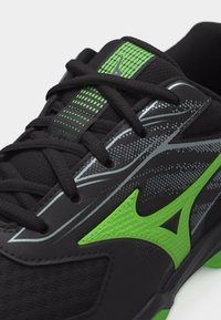 Μαύρο και πράσινο αθλητικό παπούτσι με μαύρα κορδόνια, πράσινο λογότυπο και επάνω μέρος από πλεγμένο υλικό με σχέδιο, εμφανιζόμενο σε κοντινή γωνιακή όψη.