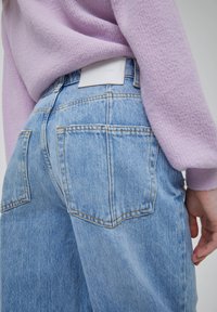 Vaqueros de mezclilla azul claro con grandes bolsillos traseros, costuras amarillas y una etiqueta blanca de la marca en la cinturilla, llevados con un suéter texturizado lavanda.