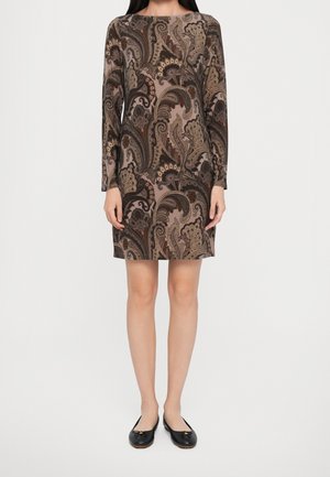 Frau trägt ein knielanges, figurbetontes Kleid mit langen Ärmeln, verziert mit einem braunen und beigen Paisley-Muster, kombiniert mit schwarzen flachen Schuhen.