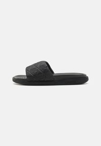 Black slide sandal made of textured rubber, featuring a wide strap with a geometric pattern and a flat sole. Lacoste logo on the side.