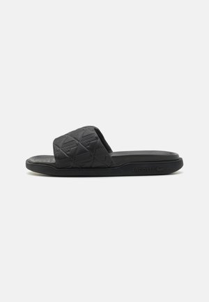Black slide sandal made of textured rubber, featuring a wide strap with a geometric pattern and a flat sole. Lacoste logo on the side.