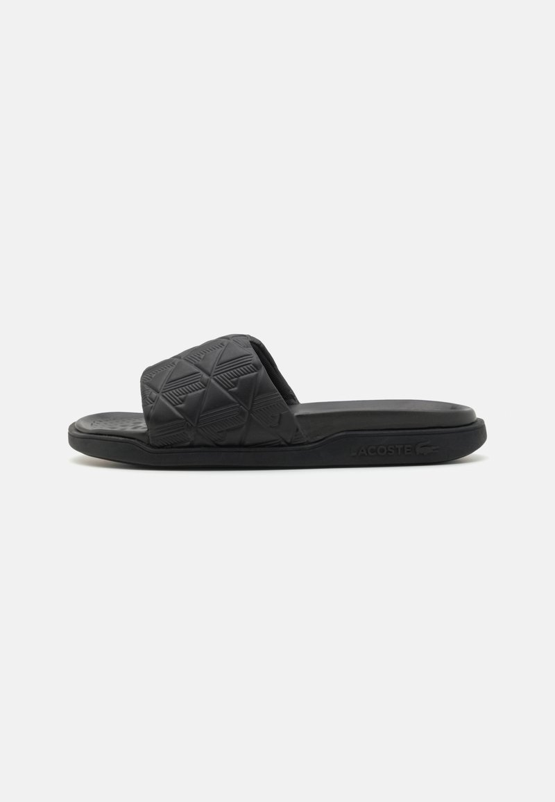Black slide sandal made of textured rubber, featuring a wide strap with a geometric pattern and a flat sole. Lacoste logo on the side.