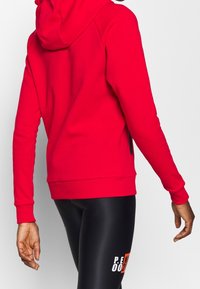 Personne portant un sweat à capuche rouge vif et un legging de sport noir avec un texte blanc et orange sur la cuisse, debout avec le dos partiellement tourné.