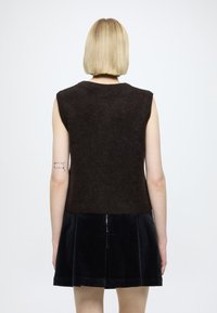 Tmavohnedý bezrukávový vlnený top s textúrovaným povrchom, v páre s čiernou manšestrovou sukňou so stredným zipsom a skladaným dizajnom.