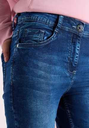 Nahaufnahme einer Person, die blaue Jeans mit sichtbaren Taschen, Nähten und Metallknopf trägt, kombiniert mit einem pinken Langarmshirt.