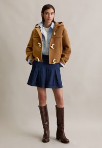 Bruine wollen dufflecoat met toggle-sluitingen, gelaagd over een lichtblauw shirt. Gecombineerd met een donkere denim plooiskirt en bruine kniehooglaarzen.