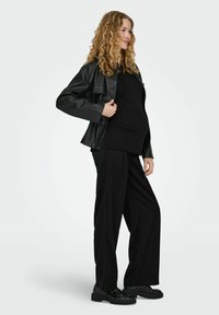 Musta tekonahkatakki mustan neuleen päällä, yhdistetty löysiin mustiin housuihin ja kiiltäviin mustiin kenkiin. Pehmeä pinta, jäsennelty muotoilu.