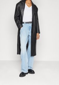 Svart läder lång kappa över en vit V-ringad tröja, i kombination med ljusblå vida jeans och svarta kraftiga stövlar.