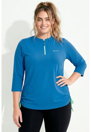 Kvinde med blond hår i en hestehale iført en blå sportstrøje med kvart lynlås og sorte leggings, smilende med hånden på hoften.
