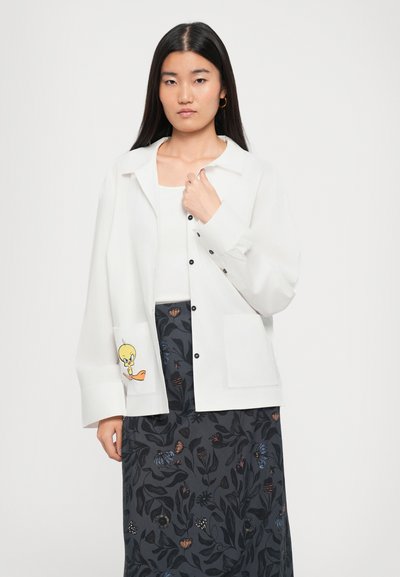 Woman wearing a white buttoned shirt with a Tweety Bird print on the pocket and a dark floral skirt, standing against a plain background.