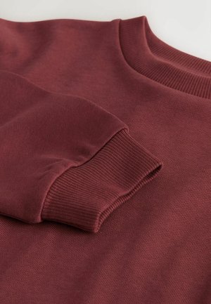 Burgundipuló, bordás nyakkal és mandzsettákkal, sima textúrájú és közepes súlyú anyagból készült. Közeli kép a ujjak részletéről.