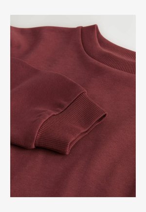 Burgundowy sweter z ryflem na dekolcie i mankietach, o gładkiej fakturze i średniej wadze materiału. Zbliżenie na detal rękawa.