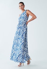 Maksi haljina plavo-bijelog zebrinog uzorka, s dizajnom na jedno rame, vezanjem u struku i glatkim materijalom. Model nosi bijele štikle.