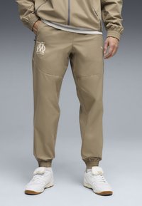 Μπεζ αθλητικό παντελόνι με στενή εφαρμογή, ελαστικές μανσέτες και λεπτομέρεια με λογότυπο. Διαθέτει λείο, ελαφρύ ύφασμα με διακριτικές τσέπες στο πλάι.
