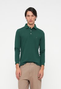 Man in een donkergroen polo met lange mouwen en lichtbruine broek staat tegen een plain witte achtergrond.