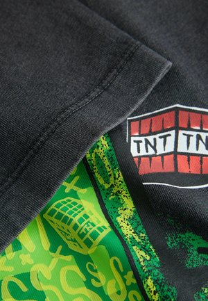 Nahaufnahme eines gefalteten dunklen Stoffes mit einem bunten Grafikdesign aus grünen und gelben Mustern sowie einem roten TNT-Block.