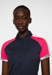 Pikétröja med mörk marinblå kropp, ärmar och krage i rosa och vitt, samt knappslå. Har "J.LINDEBERG"-logotyp på ärmen.