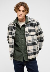 Kraciasta kurtka polarowa w kolorze kremowym, czarnym i szarym z dwoma kieszeniami na piersi, noszona na zielonej koszuli sztruksowej, zestawiona z ciemnymi dżinsami.