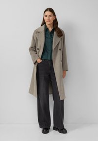 Femme portant un long manteau gris croisé, une chemise boutonnée vert foncé, un large jean noir et des chaussures noires à semelles épaisses, se tenant avec une main dans la poche.