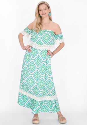 Kobieta w zielono-białej sukni maxi z odkrytymi ramionami i geometrycznymi wzorami, turkusowych kolczykach i beżowych sandałach na platformie, uśmiechająca się z rękami na biodrach.