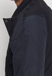 Cazadora bomber azul marino con tejido liso, cuello acanalado y acabado mate, con secciones acolchadas y botones a presión en la parte delantera.