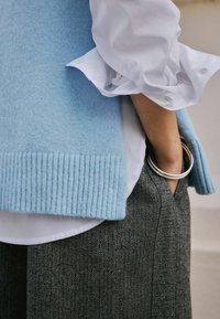 Lichtblauw gebreide vest met geribbelde zoom over een wit shirt, gecombineerd met grijs herringbone broek met textuur. Zilveren ring accent op de hand.