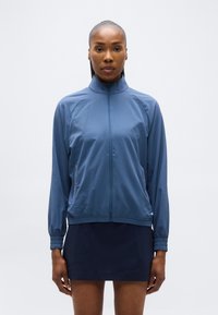 Γυναίκα με μπλε ζακέτα με φερμουάρ και σκουρόχρωμη φούστα στέκεται κοιτώντας μπροστά, μπροστά σε απλό ανοιχτό φόντο.