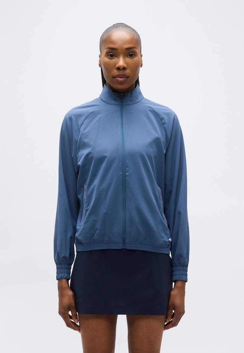 Γυναίκα με μπλε ζακέτα με φερμουάρ και σκουρόχρωμη φούστα στέκεται κοιτώντας μπροστά, μπροστά σε απλό ανοιχτό φόντο.