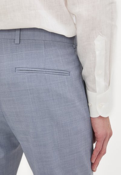 Pantaloni su misura di colore blu chiaro con una finitura testurizzata, dotati di una tasca sul retro. Abbinati a una camicia a maniche lunghe bianca con polsini con bottoni.