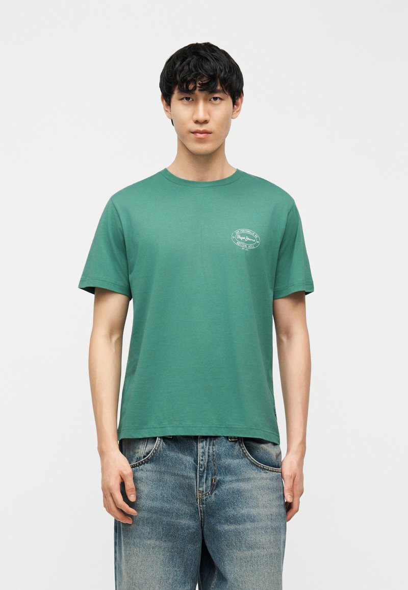 Νέος άνδρας με μαύρα μαλλιά, που φοράει πράσινο κοντομάνικο μπλουζάκι με λευκό λογότυπο στο στήθος και ξεθωριασμένο μπλε τζιν, στέκεται μπροστά σε λευκό φόντο.