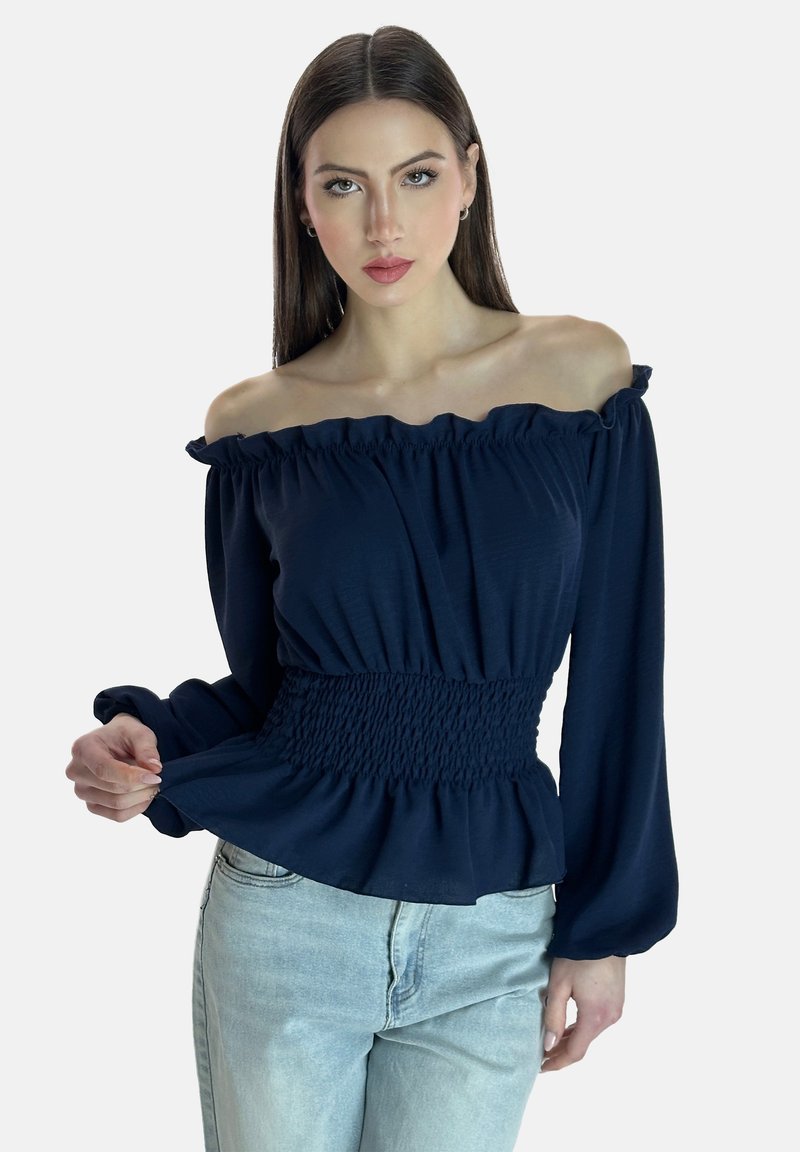 Marinblå off-shoulder blus med volangkant vid halsen, långa ärmar och resår i midjan, kombinerad med ljusblå jeans. Mjuk tygstruktur.