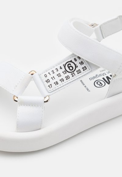 Sandal bianco con cinturini in tessuto e design con etichetta numerata. Presenta una robusta suola in gomma bianca e dettagli in hardware color oro.