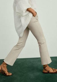 Pantalons évasés beiges en tissu doux, présentant une texture lisse et des poches latérales, accompagnés de sandales à lanières marron.