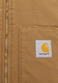 Material de pânză de culoare bej cu un fermoar auriu, având o etichetă albă pătrată cu un emblema galbenă și textul "carhartt". Detalii minime de coasere.