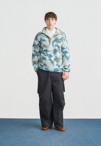 Νεαρός άνδρας στέκεται σε μπλε χαλί, φορώντας μπλε και μπεζ fleece μπουφάν με σχέδιο, λευκό πουκάμισο, φαρδιά μαύρα παντελόνια cargo και καφέ παπούτσια.