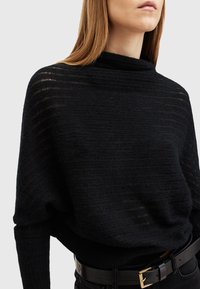 Top in maglia nera con scollatura drappeggiata alta e texture a coste. Indossato sopra una cintura nera aderente, che enfatizza il punto vita.