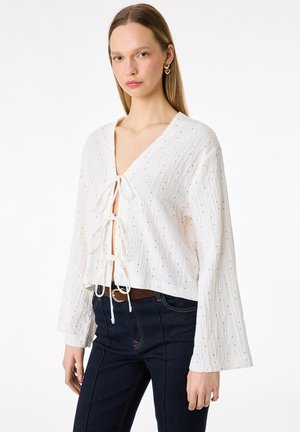 Młoda kobieta ubrana w białą bluzkę z długim rękawem w drobne kropki, z trzema wiązaniami z przodu, zestawioną z ciemnoniebieskimi dżinsami i brązowym paskiem.