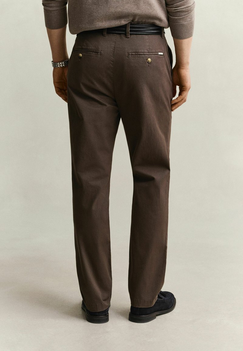 Mann trägt braune Hose, schwarzen Gürtel und schwarze Schuhe, steht mit dem Rücken zu uns, die Hände entspannt an den Seiten.