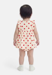 Νήπιο που στέκεται γυρισμένο ανάποδα, φοράει κρεμ φόρεμα με κόκκινη καρδιά, λευκές κάλτσες και λευκά slip-on παπούτσια σε λευκό φόντο.