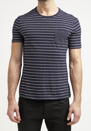Mand iført en mørkeblå kortærmet t-shirt med tynde hvide horisontale striber og en struktureret frontlomme, kombineret med sorte bukser.
