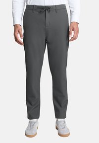 Grijze, op maat gemaakte broeken van gladde stof, met een koord in de taille, zijzakken en een slanke pasvorm. Draagt lichtgekleurde sneakers.