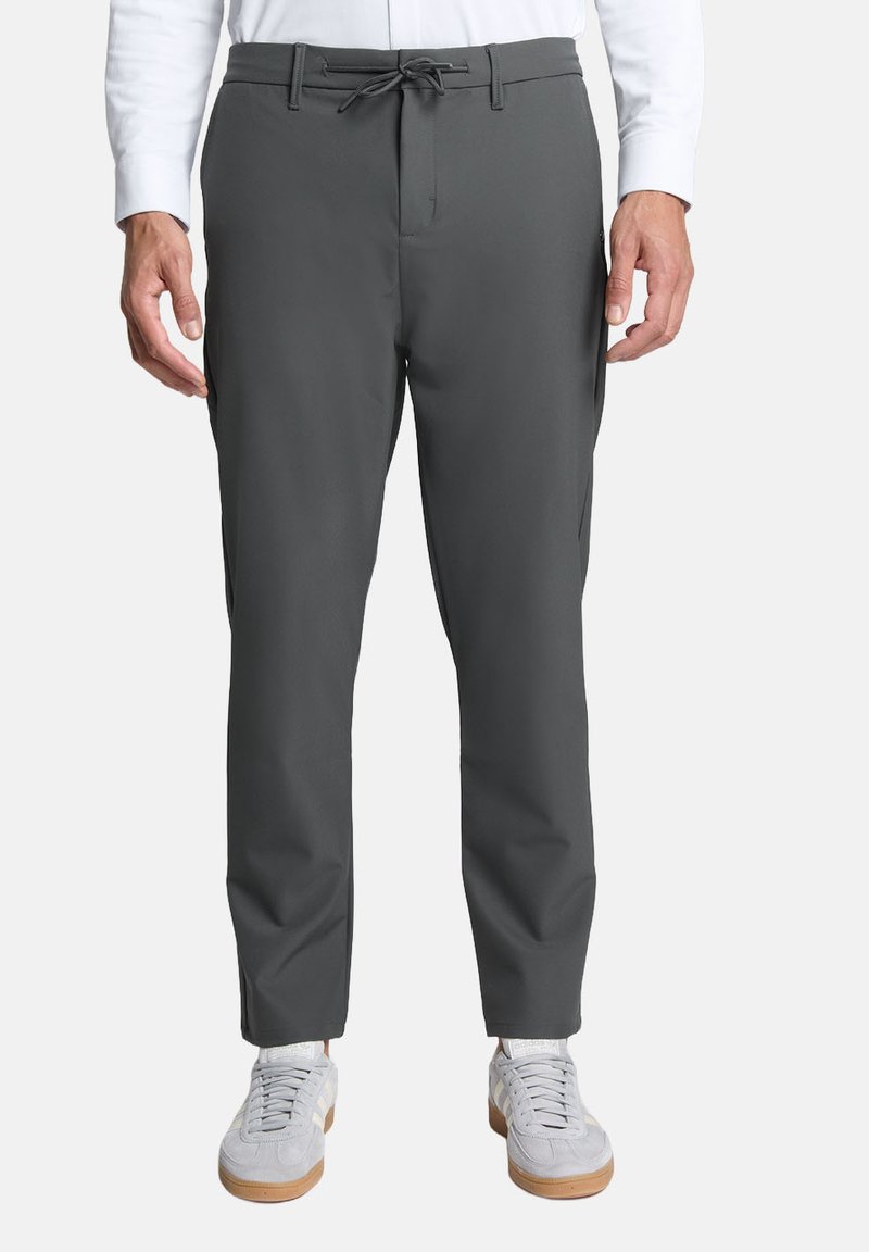 Grijze, op maat gemaakte broeken van gladde stof, met een koord in de taille, zijzakken en een slanke pasvorm. Draagt lichtgekleurde sneakers.