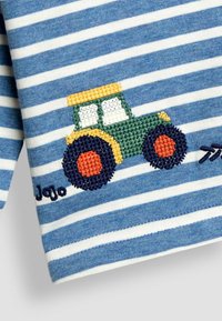 Sininen ja valkoinen raidallinen kangas esittelee brodeeratun vihreän ja keltaisen traktorin, jossa on oranssit ja laivastonsiniset pyörät, sekä nimen "Jojo."