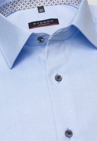 Ljusblå skjorta i slät tyg. Har en modern passform, knäppning med krage och mörka knappar med ett mönster som accent inuti kragen.