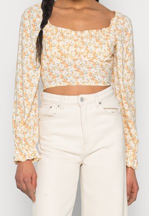 Top corto floral en crema con flores amarillas y naranjas, mangas largas abullonadas, y bordes elásticos en el cuello y las mangas. Combinado con pantalones blancos de talle alto.