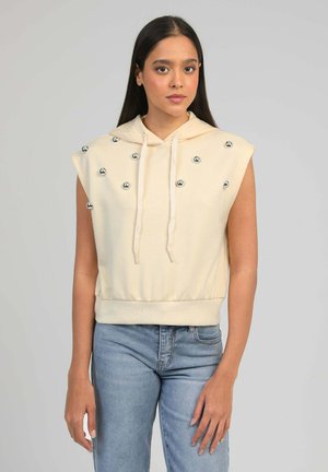 Jeune femme aux cheveux longs et foncés portant un sweat à capuche crème sans manches orné de boules argentées, assorti à un jean bleu clair.