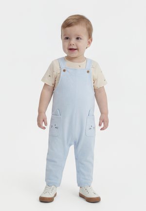 Kleinkind steht lächelnd da, trägt ein beiges Hemd und hellblaue Latzhosen mit Katzenmotiven auf den Vorderentaschen sowie weiße Turnschuhe.