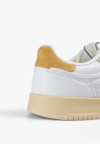 Zapatilla blanca con un tacón de ante tostado, suela de goma texturizada y simples detalles de costura. Diseñada para el uso casual y la comodidad.