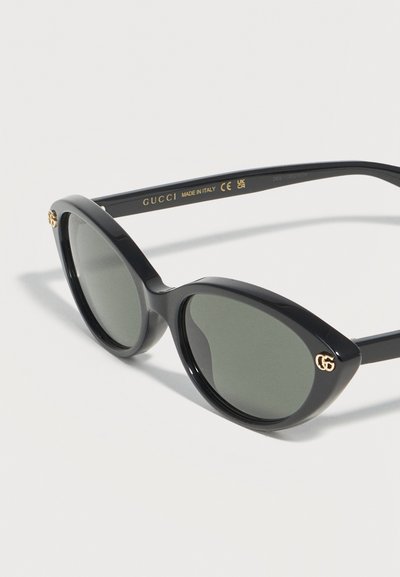 Μαύρα γυαλιά ηλίου Gucci σε σχήμα ματιού γάτας με σκούρους φακούς και χρυσά λογότυπα GG στις μπροστινές γωνίες, εμφανιζόμενα σε λευκό φόντο.